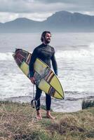 Satisfied ethnic male surfer with board smiling on grassy seasho photo