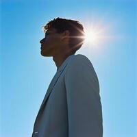 man in light suit stands against bright blue sky, with sunlight shining behind him, creating halo effect. scene conveys sense of calm and confidence photo