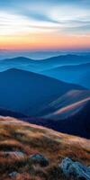 breathtaking landscape showcasing rolling mountains at sunset, with layers of blue and orange hues creating serene atmosphere. foreground features grassy hills and rocky outcrops photo