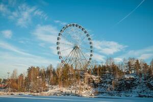 Ferris wheel standing tall against a clear blue winter sky, with snow-covered trees and rocky terrain in the foreground, creating a picturesque winter scene photo