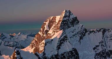 Aerial view of snow-capped peak of high mountain in rays of sunrise against clear sky video