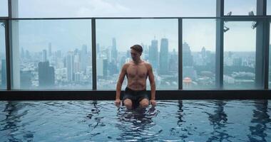 Caucasian business man relax on the edge of the roof top swimming pool in luxury hotel in big city. Young man enjoying in luxury swimming pool on the roof of skyscraper on background of city video