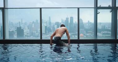 Millennial man tourist relax on the edge of the roof top swimming pool in luxury hotel in big city. Young man enjoying in luxury swimming pool on the roof of skyscraper on background of city video