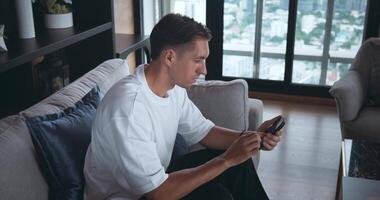 Handsome caucasian business man using mobile phone phone in living room at home, resting on sofa. Young business man browsing internet and checking videos on social networks at home office