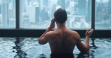 espalda ver negocio hombre hablando teléfono mientras relajarse en techo parte superior nadando piscina de rascacielos en grande ciudad. posterior ver empresario usos teléfono inteligente para negociaciones mientras relajante en piscina en ciudad antecedentes video