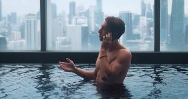 caucásico negocio hombre hablando teléfono mientras relajarse en techo parte superior nadando piscina de rascacielos en grande ciudad. Rico negocio masculino usos teléfono inteligente para negociaciones mientras relajante en el piscina en ciudad antecedentes video