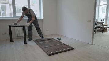 A builder in a work overalls is assembling a table. Assembling furniture in a new apartment. video