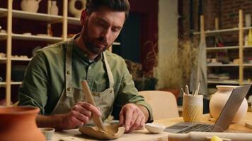 Caucasian male man thoughtful creative small business owner businessman guy potter craftsman making stoneware tool watching laptop tutorial pottery lesson online computer device indoors studio video