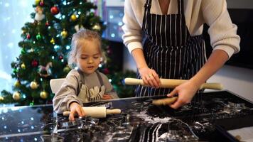 Little daughter sits next to her mother rolling out the dough on the table with a rolling pin video