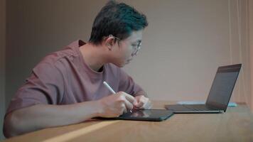Focused designer a young man meticulously works on his tablet using a stylus, his concentration evident as he collaborates with a laptop. video
