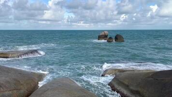 Scenic coastal landscape featuring large rocky formations by the ocean. The turquoise sea waves crash against the rocks under a vibrant blue sky with scattered white clouds video