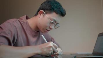 Focused design a young man wearing glasses concentrates intently on his digital tablet, a pen in hand, as he works on a creative project. video