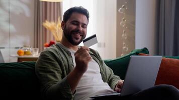 Happy smiling Caucasian man male guy shopper client customer homeowner using laptop typing computer holding credit card bank paying online ecommerce shopping internet buy transaction indoor home room video