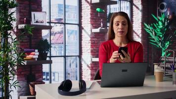 Focused woman in a home office loft talking on a mobile phone while working on laptop. She juggles calls, meetings and inquiries, using communication skills and creativity to solve tasks. Camera B. video