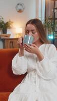 Smiling young woman sitting on sofa drinking cup of warm coffee or herbal tea in morning at home video