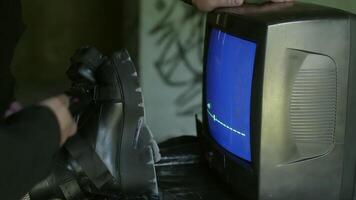 Hands of a man holding a remote, ready to control a flickering TV. Woman with crossed legs in heels and fishnet stockings rest nearby. They sit in a graffiti-filled, abandoned room video