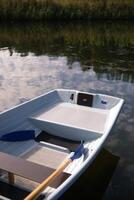 A close-up of a white rowboat with wooden oars floating on a calm river, reflecting the sky and surrounded by reeds. photo
