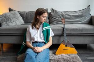 Thoughtful female musician sit on floor, observe balalaika. Challenge of mastering traditional music photo