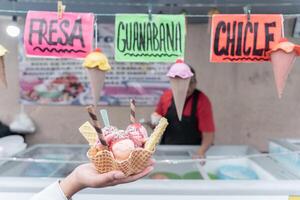 A hand is holding a traditional Mexican decorated ice cream in front of a street market stall with flavor signs photo