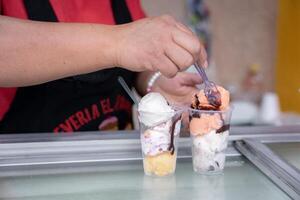 An Hispanic adult woman is serving some ice cream balls with a spoon in plastic cups in the street photo