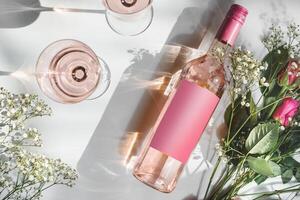 Rose wine bottle with empty pink mock up label, two wine glasses and romantic flower bouquet on white background with sunlight and shadow. Modern wine concept. Top view photo