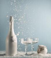 White champagne bottle and glasses with Christmas bauble on table with snowfall at blue wall background. New years concept with drink and falling snow. Front view. photo