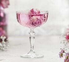 Close up of sparkling pink drink with rose petals in champagne glass at white table with blurred background. Aperitif with elegant decoration for celebration. Front view. photo