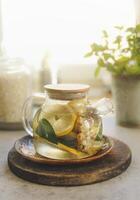 Glass teapot with lemon, mint and ginger on wooden cutting board at window background with natural light. Healthy tea with vitamin c in cold time. Front view. photo