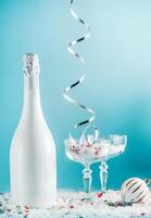 Christmas and New Year setting with champagne bottle, glasses, falling silver ribbon, snow and bauble and at light blue background. Front view photo