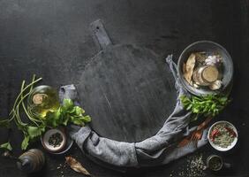 Kitchen flavor and seasoning ingredients with kitchen utensils around empty black cutting board. Top view. Frame photo