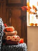 Autumn still life with folded cozy sweaters, wool blanket, pumpkin, candle at window background with sunlight. Warm clothes and seasonal interior decoration at home. Front view photo