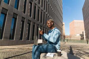 Self employed young ethnic guy using smartphone during work on netbook on street photo