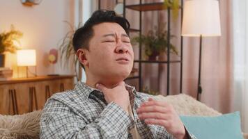 Upset Chinese young man holds hands over sore throat with painful neck and frowning on sofa at home video