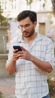 Hispanic young man using smartphone text messages browsing internet smiling at camera outdoors video