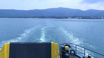 Scenic view from the back of a ferry leaving the shore, creating rippling waves in the blue sea with a mountain landscape in the background video