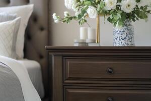 A close-up view of a stylish Transitional interior design style bedside table featuring a vase of white flowers, candles, and a textured bed in the background. photo