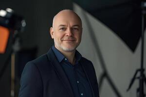 A confident middle-aged sales manager man with a short beard, wearing a dark suit and blue shirt, smiles warmly at the camera. The background is softly blurred, with studio lighting equipment visible. photo