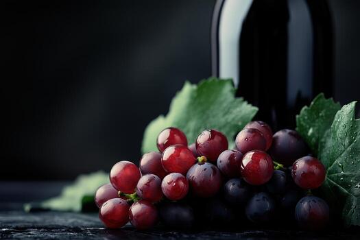 A close-up of fresh red and black grapes with green leaves, set against a dark background featuring a wine bottle. photo