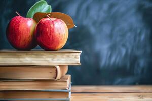 Two red apples on top of a stack of books, with a green leaf resting on the apples. The background is a blurred blackboard, suggesting an educational setting. photo
