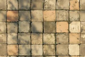 Aerial view of a textured pavement made of square tiles in various shades of beige and gray, some tiles showing signs of wear and moss growth. photo