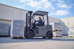 A worker operating a forklift in a warehouse, maneuvering around stacked pallets. The scene is bright with blue skies and industrial buildings in the background. photo