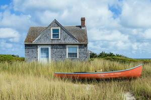 A quaint wooden cape cod house with a gray shingle exterior sits amidst tall grass, with a bright orange canoe resting nearby. The sky is partly cloudy, creating a serene coastal atmosphere. photo