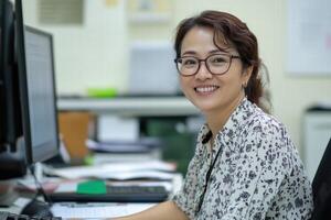 A woman in glasses is smiling while sitting at a desk photo