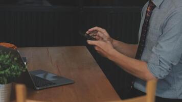 A man in a shirt and tie sitting at a table with a laptop and a phone video