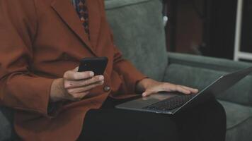 A man in a suit sitting on a couch with a laptop and cell phone video