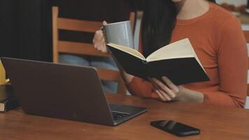 A woman is reading a book while sitting at a table with a laptop video