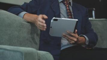 A man in a suit and tie sitting on a couch with a tablet video
