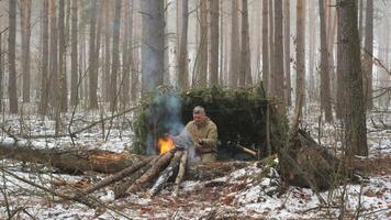en man värms upp hans händer nära en brand i främre av en handgjort tall-gren skydd i en snöig skog. en Sann vildmark överlevnad och utomhus- äventyr scen. video