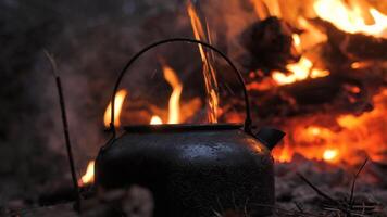 A rustic black kettle heats over an open fire, surrounded by glowing embers. A perfect outdoor survival and bushcraft scene in the wilderness. video