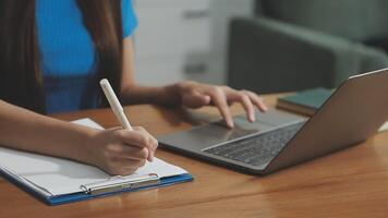 A woman is writing on a notebook while using a laptop video